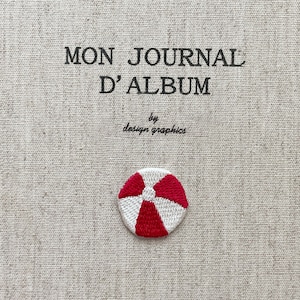 May include: Embroidered journal cover with the text "MON JOURNAL D'ALBUM" and "by design graphics." A small, red and white beach ball design is stitched on the cover. The background is a textured, beige fabric.
