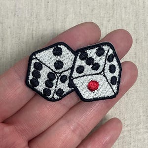 Puede incluir: Parche bordado con dos dados. Los dados son blancos con puntos negros y un borde negro. Un dado tiene un solo punto rojo. El parche se sostiene en una mano.