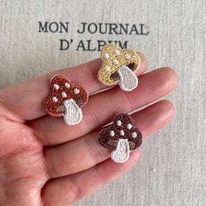 Könnte beinhalten: Drei kleine gestickte Pilzaufnäher in den Farben Rostorange, Goldgelb und Dunkelbraun. Jeder Aufnäher hat einen weißen Stiel und einen Hut, der mit kleinen weißen Punkten verziert ist. Die Stickerei weist eine sichtbare Fadenstruktur auf.
