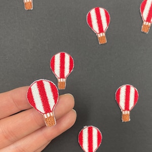 May include: Multiple embroidered patches shaped like hot air balloons. Each patch features vertical red and white stripes on the balloon canopy and a brown woven basket at the base. The patches are arranged in a scattered pattern across the frame.
