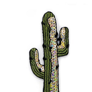 Könnte beinhalten: Ein grüner, kaktusförmiger Aufnäher mit braunem Sockel und schwarzer Kontur. Der Kaktus ist mit schillernden Pailletten in der Mitte und an den Armen verziert. Der Aufnäher ist zum Aufnähen oder Aufbügeln auf Stoff gedacht.