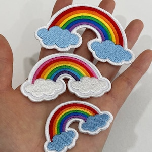 Op de afbeelding: Drie geborduurde regenboogpatches met een witte rand en blauwe wolkenaccenten. De regenbogen hebben de kleuren rood, oranje, geel, groen, blauw en paars. De patches zijn in verschillende maten.