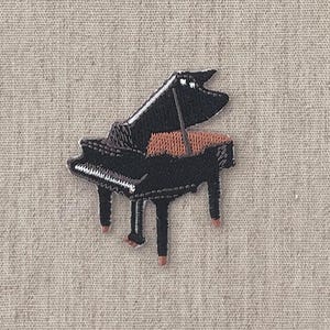 Puede incluir: Una portada de diario beige con el texto "MON JOURNAL D'ALBUM" y "by design graphics" en negro. Un piano de cola bordado en negro y naranja en la parte inferior. El diario tiene una apariencia de tela texturizada.