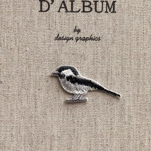 Peut inclure: Une couverture de journal avec le texte "MON JOURNAL D'ALBUM" et "by design graphics" en noir. Un petit oiseau brodé en noir, blanc et gris est sur la couverture. La couverture est en tissu beige texturé.