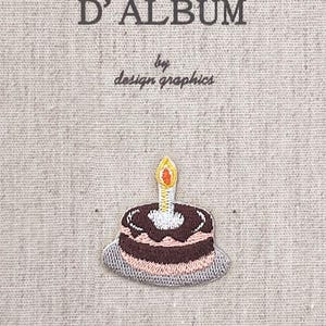 Può includere: Una copertina di diario in tessuto con la scritta "MON JOURNAL D'ALBUM" e "by design graphics". Una piccola torta ricamata con una candela accesa è centrata in basso. Lo sfondo è un tessuto beige chiaro.