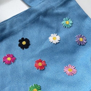 Pode incluir: Uma sacola azul clara decorada com flores de margarida bordadas em várias cores, incluindo rosa, branco, preto e verde. As margaridas têm centros amarelos. A sacola tem duas alças.