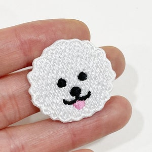 May include: A white embroidered patch in the shape of a dog's face. The dog has black eyes and nose, and a pink tongue. The patch has a scalloped edge.