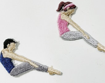 May include: Two embroidered patches depicting figures in a seated yoga pose. One figure wears a blue top, the other a pink top. Both have gray pants and are embroidered with various colors. The patches are on a white background.