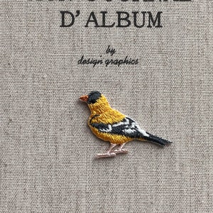 May include: A beige journal cover with the text "MON JOURNAL D'ALBUM" and "by design graphics" in black. A yellow, black, and white embroidered bird is on the cover. The journal cover has a textured appearance.