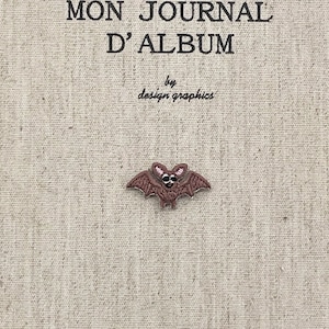May include: A beige journal cover with the French text "MON JOURNAL D'ALBUM" and "by design graphics". A small, brown bat-shaped embellishment is centered below the text. The bat has a skull-like face.