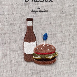 May include: Embroidered journal cover with the text "MON JOURNAL D'ALBUM" and "by design graphics". Features a brown bottle with a white label and a burger with a blue toothpick on a beige fabric background.