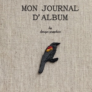Puede incluir: Una portada de diario con el texto "MON JOURNAL D'ALBUM" y "by design graphics". Un pájaro negro con detalles rojos y amarillos está bordado en la portada. El fondo es una tela beige texturizada.