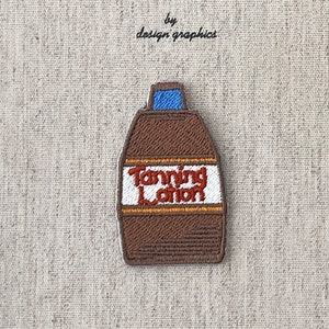 Op de afbeelding: Geborduurde patch met een bruine zonnebrandlotionfles met de witte tekst "Tanning Lotion". De patch is geplaatst tegen een beige stoffen achtergrond. Boven de patch staat de tekst "MON JOURNAL D'ALBUM" en "by design graphics".
