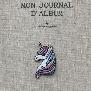 Puede incluir: Parche de unicornio bordado con cara blanca, melena rosa y morada, y cuerno azul y rosa. El parche está sobre una tela beige con el texto "MON JOURNAL D'ALBUM" y "by design graphics".