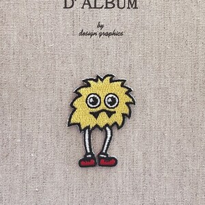 Könnte beinhalten: Ein gelber Monsteraufnäher mit schwarzen Umrissen, weißen Augen und roten Schuhen. Der Aufnäher befindet sich auf einem beigefarbenen Hintergrund mit dem Text "MON JOURNAL D'ALBUM" und "by design graphics" in Schwarz.