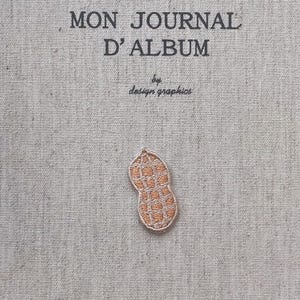 Può includere: Una copertina di diario in tessuto beige con la scritta "MON JOURNAL D'ALBUM" e "by design graphics" stampata in nero. Una piccola toppa ricamata a forma di arachide è attaccata alla copertina.