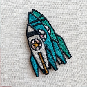 May include: Three embroidered rocket patches in teal, white, and green. Each patch features a star design with a gold centre. The rockets have black outlines and are arranged diagonally on a neutral fabric background.