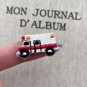 May include: Embroidered patch of a white ambulance with red and black details. The ambulance has orange lights on top and black wheels. The patch is small and detailed, perfect for adding to clothing or accessories.