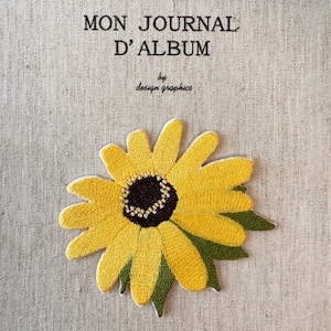 May include: Embroidered journal cover with the text "MON JOURNAL D'ALBUM" and "by design graphics." The cover features a large, yellow sunflower with a dark brown center and green leaves. The background is a light beige fabric.