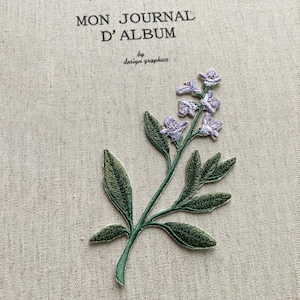 May include: Embroidered floral patch with a sprig of lavender flowers and green leaves. The patch is on a beige fabric with the text "MON JOURNAL D'ALBUM" and "by design graphics".