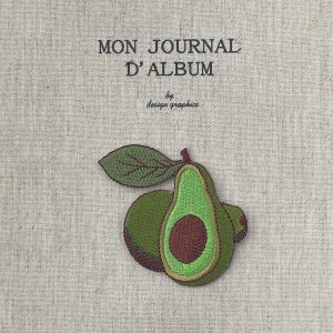 May include: A beige fabric journal cover with the text "MON JOURNAL D'ALBUM" and "by design graphics" in black. An embroidered avocado with a leaf is in the lower center. The avocado is green with a brown pit and a dark purple outline.