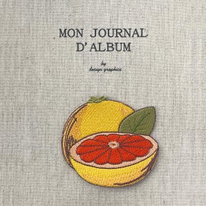 Puede incluir: Cubierta de diario bordada con el texto "MON JOURNAL D'ALBUM" y "by design graphics". La cubierta presenta un pomelo amarillo con interior rojo y una hoja verde. El fondo es una tela gris claro.