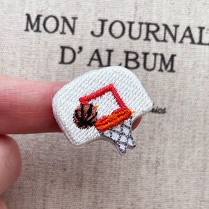 Könnte beinhalten: Gestickter Aufnäher mit einem Basketballkorb und einem Ball. Das Brett ist weiß, der Ring ist rot und orange, und das Netz ist grau. Der Basketball ist braun und schwarz. Im Hintergrund steht der Text "MON JOURNAL D'ALBUM".
