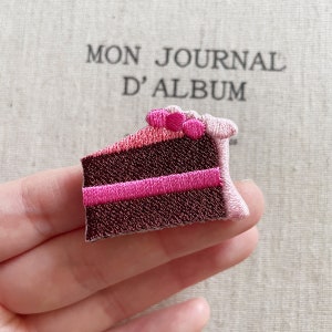 May include: Embroidered patch depicting a slice of chocolate cake with pink frosting and pink dot decorations. The patch is held in a hand, with the text "MON JOURNAL D'ALBUM" visible in the background.