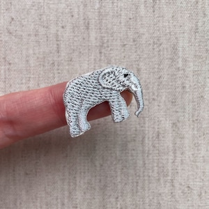 Op de afbeelding: Een kleine, geborduurde patch in de vorm van een olifant. De patch is zilverkleurig met zwarte ogen en wordt door een vinger vastgehouden. De olifant heeft een gedetailleerd, getextureerd uiterlijk.