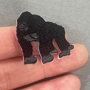 Könnte beinhalten: Ein schwarzer Gorilla-Aufnäher mit weißem Rand. Der Aufnäher ist mit einem detaillierten Design bestickt, das das Fell und die Form des Gorillas zeigt. Der Aufnäher ist wahrscheinlich für Kleidung oder Accessoires bestimmt.