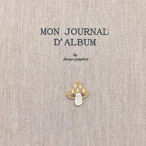 May include: A beige journal cover with the text "MON JOURNAL D'ALBUM" and "by design graphics" in black. A small embroidered mushroom with a tan cap and white stem is in the lower right corner.