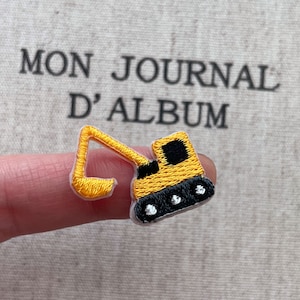 Könnte beinhalten: Gestickter Aufnäher einer gelb-schwarzen Baggers mit weißem Rand. Der Aufnäher wird vor einem Hintergrund mit dem Text "MON JOURNAL D'ALBUM" gehalten. Der Bagger hat ein detailliertes Design.