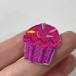 May include: Embroidered cupcake patch with pink frosting and a purple base. The frosting is decorated with yellow and purple sprinkles. A fun accessory for clothing or bags.