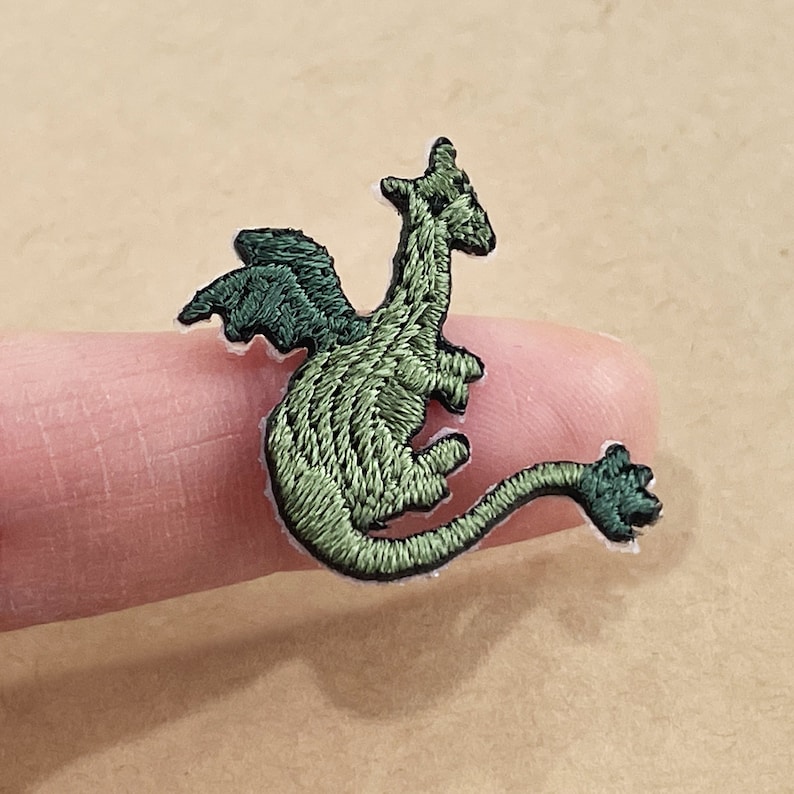 以下が含まれることがあります： ダークグリーンの翼と長い尾を持つ緑色のドラゴンの刺繍パッチ。 ドラゴンは、テクスチャードステッチと黒いアウトラインで詳細に描かれています。 パッチは白い裏地の上にあります。