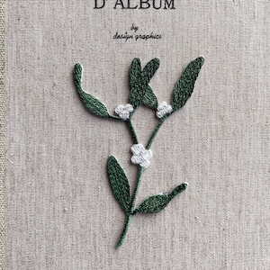 以下が含まれることがあります： ベージュ色の生地に、緑の葉と白い実の刺繍が施されたヤドリギのデザイン。上部には「D' ALBUM」と「by design graphics」の文字が見えます。刺繍は細かく、質感があります。
