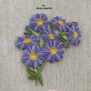 Pode incluir: Buquê bordado de flores roxas com centros amarelos e caules e folhas verdes. O texto "by design graphics" está no canto superior esquerdo. As flores estão em um tecido bege claro.