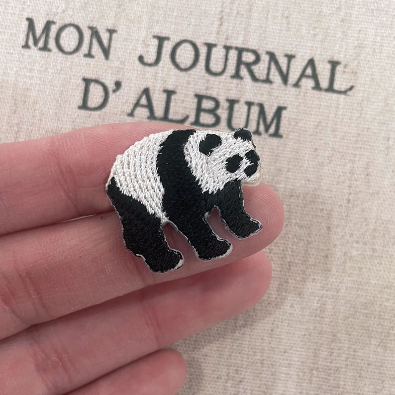May include: Embroidered panda bear patch in black and white. The patch is in the shape of a panda bear standing on all fours. The background has the text "MON JOURNAL D'ALBUM".