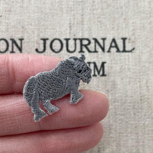 以下が含まれることがあります： グレーの刺繍されたサイのパッチ。サイは歩いているポーズで描かれており、詳細なステッチがテクスチャの外観を作り出しています。パッチは指の間に保持され、背景には「JOURNAL」という単語が部分的に表示されています。