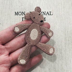 May include: Embroidered brown teddy bear patch with a cream-colored belly and paw pads. The bear has a black nose and is in a playful pose. The patch is a cute addition to clothing or accessories.