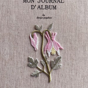 May include: Embroidered floral design featuring pink and white flowers with green leaves and stems. The text "MON JOURNAL D'ALBUM" and "design graphics" are visible on the pink fabric background.