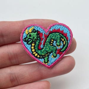 May include: Embroidered heart-shaped patch featuring a green dragon with yellow horns and accents. The dragon is set against a light blue background, framed by a pink border. The patch is likely for clothing or accessories.