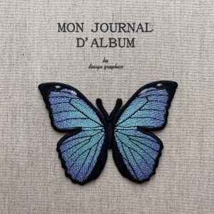 May include: Embroidered butterfly patch with iridescent blue and teal wings, outlined in black. The patch is on a beige fabric background. Text at the top reads "MON JOURNAL D'ALBUM" and "by design graphics" below.