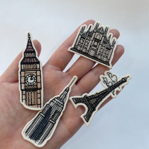 May include: Embroidered patches featuring iconic landmarks: Big Ben, the Empire State Building, the Eiffel Tower, and a cathedral. Each patch has a cream-colored border and detailed stitching in shades of brown and black.