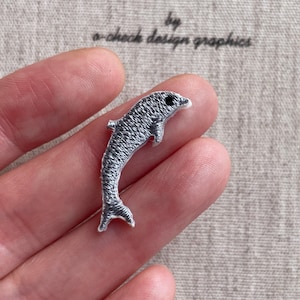 以下が含まれることがあります： グレーと白の刺繍入りイルカのパッチ。イルカは湾曲した体と小さな黒い目を持っています。パッチは手で持たれています。「by a-check design graphics」というテキストが背景にあります。