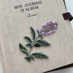 Op de afbeelding: Een beige linnen journal met de tekst "MON JOURNAL D'ALBUM" en "by design graphics". Het tijdschrift is versierd met een geborduurde tak met groene bladeren en paarse bloemen. Een bruin lint is aan het tijdschrift bevestigd.