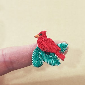 Puede incluir: Pájaro cardenal rojo bordado posado en una rama bordada verde y blanca. El cardenal tiene una textura detallada, con un pequeño pico amarillo y detalles en negro. La rama tiene una textura detallada. El tema general es la naturaleza y las aves.