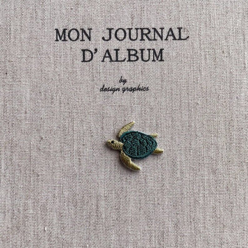 May include: A beige journal cover with the text "MON JOURNAL D'ALBUM" and "by design graphics" in black. A small embroidered sea turtle with a teal shell and tan flippers is on the cover.