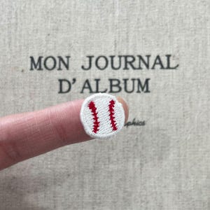 Op de afbeelding: Een kleine, geborduurde baseball patch met rode stiksels op een witte achtergrond. De patch wordt voor een achtergrond met de tekst "MON JOURNAL D'ALBUM" gehouden.