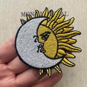 May include: Embroidered patch featuring a celestial design of a sun and moon. The sun is yellow with detailed rays, and the moon is a light gray with a face and script. The patch has a black border.