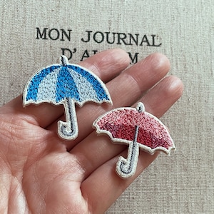 Puede incluir: Dos parches bordados con forma de paraguas. Uno es azul y blanco, el otro es rojo. Ambos tienen un borde blanco y un asa curvada. El fondo tiene el texto "MON JOURNAL D'ALBUM".